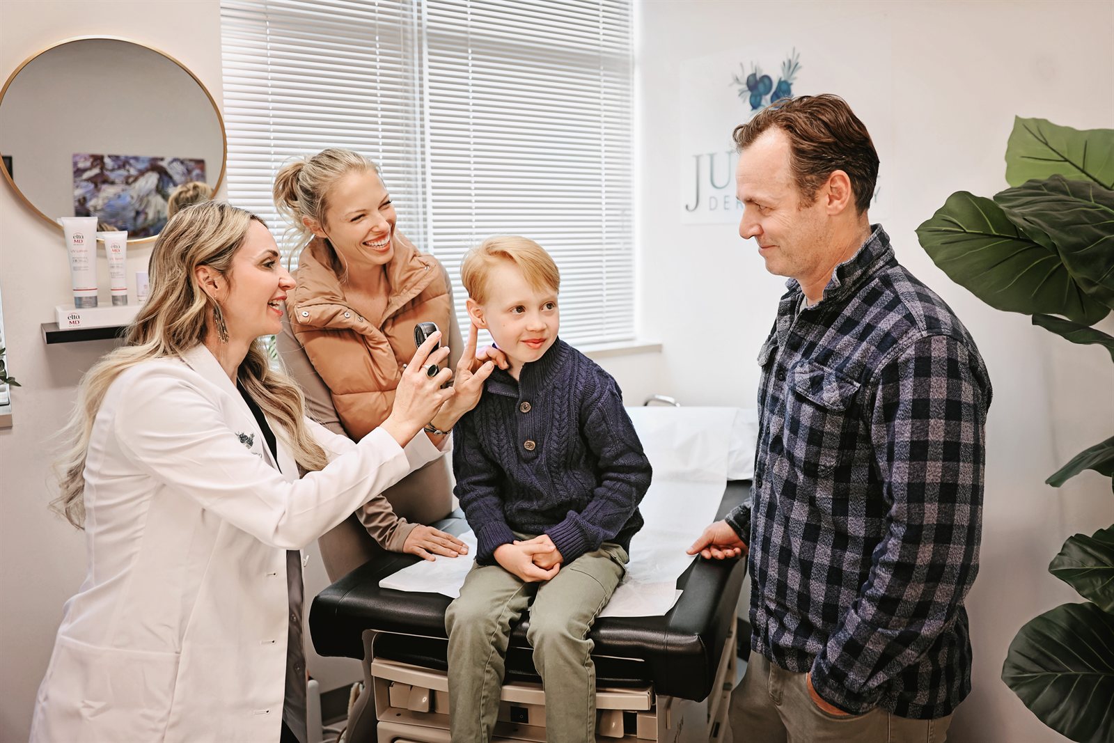 Dr. Karla Pivik examining a young patient's skin with a dermatoscope while his family looks on at Juniper Dermatology